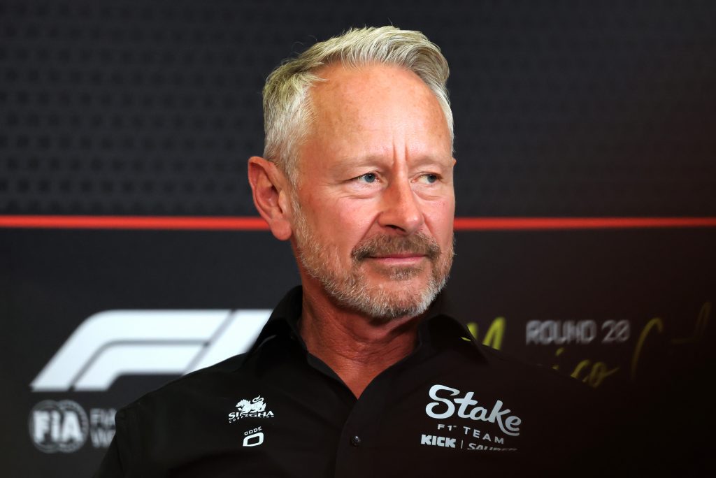 Close-up portrait of a smiling, gray-haired man with a trimmed beard wearing a black shirt with sponsor logos, looking to the right.