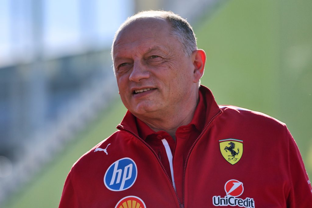 Older man in a red Ferrari jacket outdoors, looking to the left with a slight smile in daylight.
