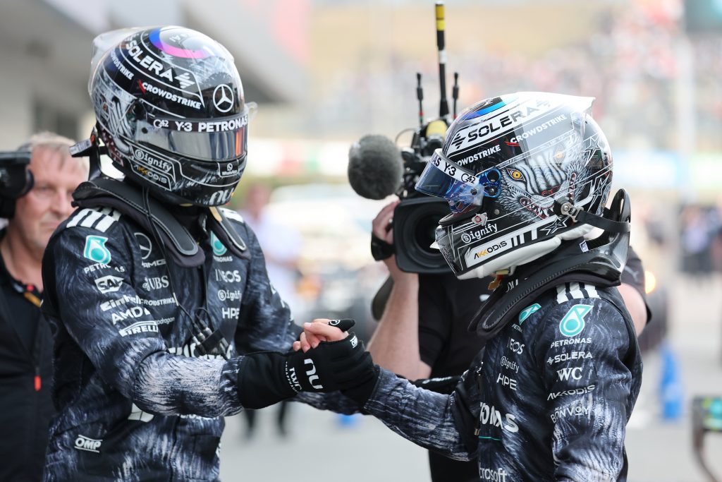 Two race drivers wearing detailed helmets and dark sponsor-covered suits shake hands on a pit-lane, with a camera crew in the background.