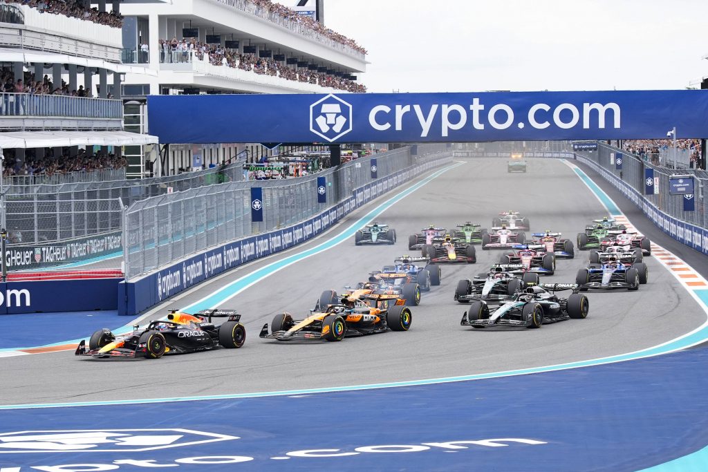 Formula 1 cars launch from the grid on a street circuit as crowds watch from grandstands under a large crypto.com banner.