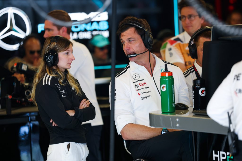 Two Mercedes AMG team members in a pit area wearing headsets; a woman stands with arms crossed while a man sits at a laptop nearby.
