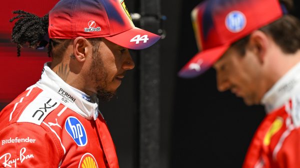 Two Ferrari drivers in red racing suits face each other; one wears a cap with #44 and sponsorship logos like Shell and HP.