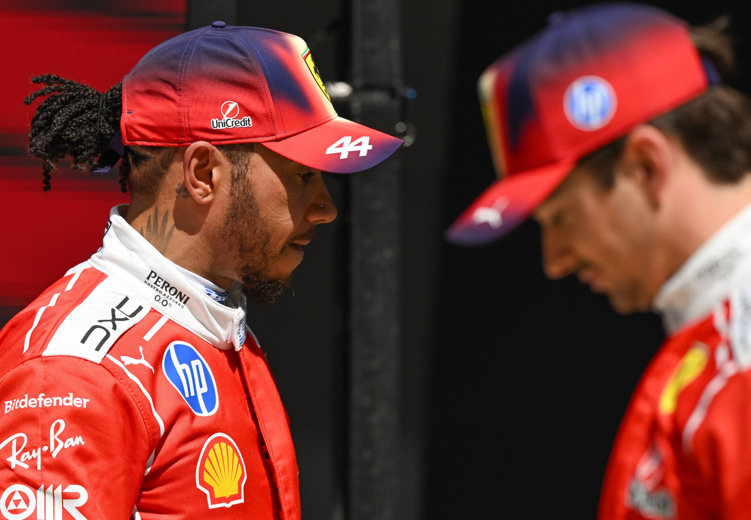 Two Ferrari drivers in red racing suits face each other; one wears a cap with #44 and sponsorship logos like Shell and HP.