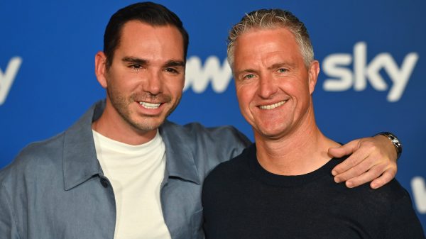 Two men smiling and posing with an arm around each other in front of a blue backdrop with partial white text.