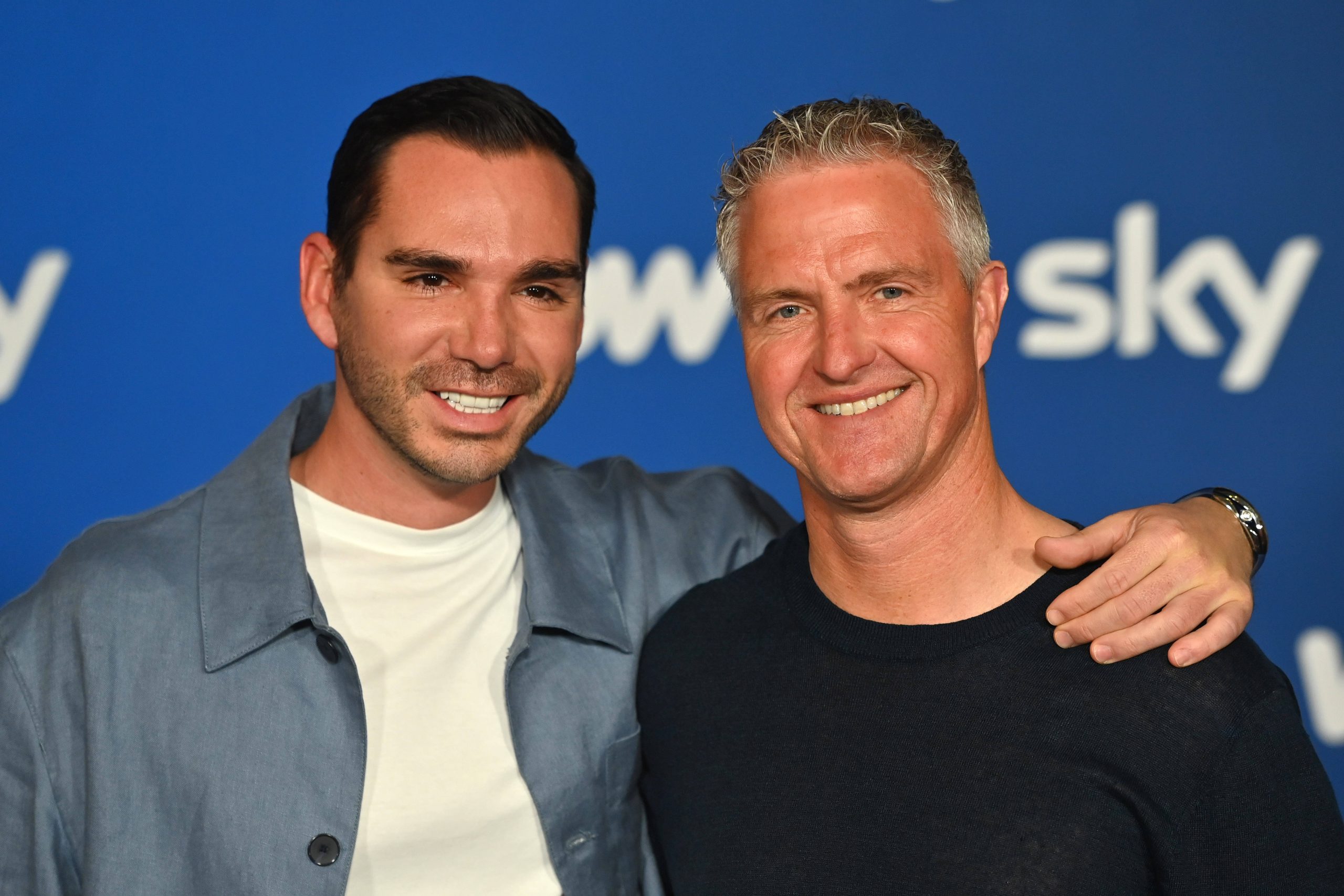 Two men smiling and posing with an arm around each other in front of a blue backdrop with partial white text.