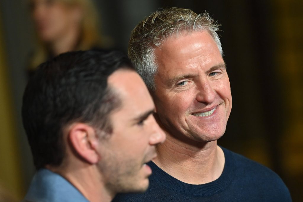 Two men at an event: one in the foreground talking, the other facing the camera with a friendly smile.